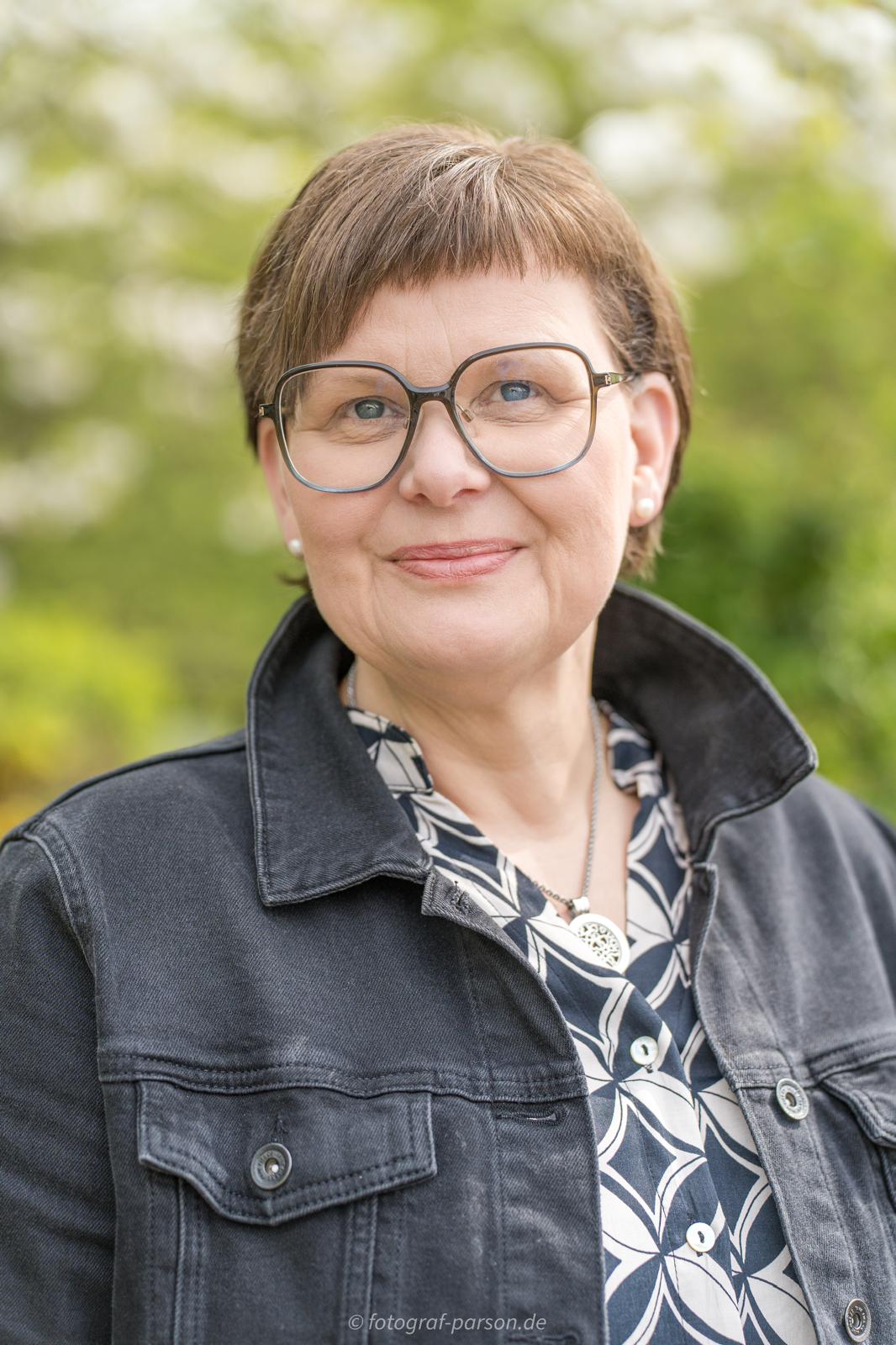 Portrait of a smiling woman with short brown hair and large round glasses, wearing a dark denim jacket outdoors.